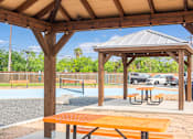 Thumbnail 3 of 11 - A wooden pavilion with picnic tables is situated in a park with a playground in the background.