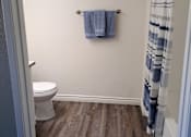 Thumbnail 16 of 36 - A bathroom with a toilet, a towel, and a shower curtain at Raintree Apartments, CA 92346