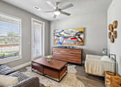 Thumbnail 5 of 36 - A living room with a grey couch, a coffee table, a painting, a fan, and a window with blinds.
