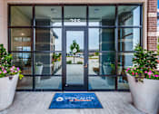 Thumbnail 16 of 36 - Glass-front building entry with covered walkway and planters at Vercanta at Burleson.