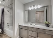 Thumbnail 9 of 35 - A bathroom with a white toilet, a bathtub with a shower curtain, a sink with a mirror and lights above it, and a potted plant on the counter.