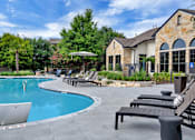 Thumbnail 20 of 35 - Pool deck with chaise lounges beside a stone-style clubhouse exterior and gardens.