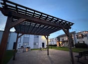Thumbnail 5 of 27 - A wooden pergola structure is in the foreground of a park-like setting with benches and a building in the background.