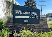 Thumbnail 2 of 14 - A sign for Whispering Oaks Apartments stands in front of a tree.