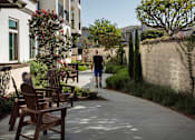 Thumbnail 57 of 114 - A man walks down a sidewalk past a row of wooden chairs.