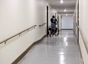 Thumbnail 90 of 103 - A man in a black shirt and blue shorts is pushing a wheelchair down a long hallway with white walls and a reflective floor.