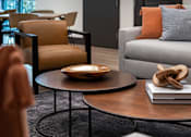 Thumbnail 89 of 90 - A living room with a grey couch, a brown chair, and a round table with a book and a pretzel on it.
