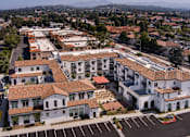Thumbnail 63 of 90 - A large white building with a red roof is surrounded by other buildings and a parking lot.