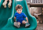 Thumbnail 11 of 21 - A young boy in a blue shirt and shorts is sitting on a green slide.