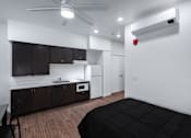 Thumbnail 119 of 127 - A black bed is in a white room with a kitchen area in the background.