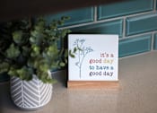 Thumbnail 36 of 127 - A card with the text "it's a good day to have a good day" is displayed in front of a plant.