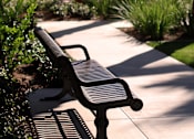 Thumbnail 13 of 45 - A park bench sits on a concrete path with green plants and trees in the background.