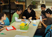 Thumbnail 8 of 126 - A woman is helping a young girl with her schoolwork at a table with other children.
