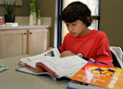 Thumbnail 43 of 126 - A young boy in a red shirt is reading a book at a table.