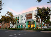 Thumbnail 4 of 16 - A modern building with a white facade and green accents is surrounded by trees and parked cars.
