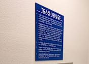 Thumbnail 91 of 102 - A blue sign with white text titled Trash Rules is on a wall above two silver trash receptacles.