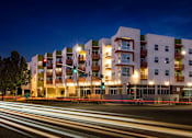 Thumbnail 65 of 94 - A modern apartment building is lit up at night with motion blurred street lights in the foreground.
