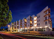 Thumbnail 69 of 94 - A modern apartment building is lit up at night with cars passing by on the street.