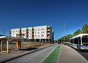 Thumbnail 46 of 94 - A bus is driving on a road with a bus stop on the left.