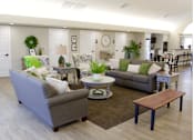 Thumbnail 4 of 20 - A living room with a grey couch, a green cushion, a white coffee table, and a brown bench.