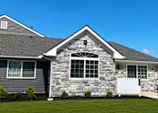 Thumbnail 6 of 42 - A house with a grey siding and a stone facade with a white fence in front.