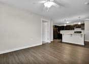 Thumbnail 9 of 25 - A room with a ceiling fan and a kitchen area in the background.