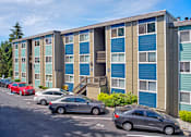 Thumbnail 9 of 14 - Community apartments painted shades of blue and beige with parked cars along the landscaped path.