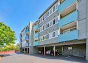 Thumbnail 11 of 14 - Blue, teal, and beige apartment building with carports underneath. Green trees line the left side of the paved driveway.