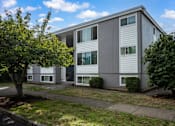 Thumbnail 1 of 22 - A grey building with white trim and windows is surrounded by greenery.