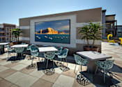 Thumbnail 11 of 57 - A large screen is mounted on a wall and surrounded by chairs and tables at Purl Apartments, San Diego, 92108