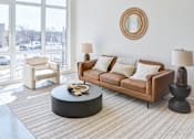 Thumbnail 10 of 19 - A living room with a brown couch, a white chair, and a round table with a yellow object on it.