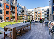 Thumbnail 37 of 61 - A wooden table with chairs is in the foreground of a patio area with a building in the background.