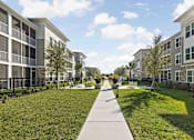Thumbnail 26 of 61 - A walkway in the middle of a grassy area with apartment buildings on either side at Verity Luxury Apartments, Vero Beach, FL 32960