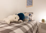 Thumbnail 26 of 36 - A white teddy bear is lying on a bed with a grey and white checkered comforter.