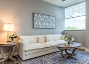 Thumbnail 6 of 33 - A living room with a white couch, a round table with a vase of flowers, and a framed picture on the wall.
