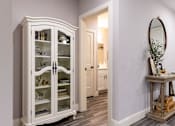 Thumbnail 48 of 50 - A white china cabinet with glass doors stands in a room with a table and mirror.