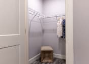 Thumbnail 42 of 50 - A small closet with clothes hanging on a wire rack and a stool in front.