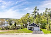 Thumbnail 25 of 60 - A house with a garage is situated on a grassy area with trees and a body of water in the background.