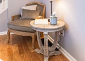 Thumbnail 10 of 53 - A table with a lamp and a book on it with a chair and a painting in the background.