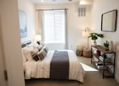 Thumbnail 9 of 34 - A bedroom with a bed, a window with blinds, a ceiling fan, a mirror, and a shelf with a plant and other items.