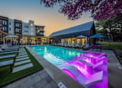 Thumbnail 1 of 30 - A resort-style pool surrounded by lounge chairs and umbrellas near a community gym and apartment buildings.