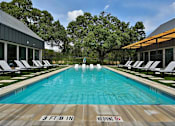 Thumbnail 7 of 30 - A gated swimming pool lined with lounge chairs near a clubhouse and large trees.