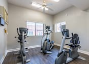 Thumbnail 10 of 13 - A room with a white ceiling and a fan, containing a treadmill, a stationary bike, and a rowing machine.