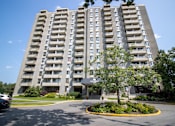 Thumbnail 2 of 11 - A large grey apartment building with a parking lot in front.