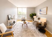 Thumbnail 4 of 11 - A living room with a grey rug, a grey sofa, a wooden chair, a wooden coffee table, a white side table, a white wall, a window, a plant, and a painting.