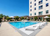 Thumbnail 10 of 27 - A large swimming pool with lounge chairs and umbrellas in front of a tall building.