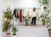 Thumbnail 15 of 17 - A woman is looking at clothes on a rack in a room with potted plants.