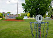 Thumbnail 98 of 98 - A disc golf basket with the number 18 on it is in the foreground of a grassy area with a sign that says Lions Park in the background.