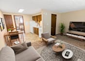 Thumbnail 4 of 12 - A living room with a grey couch, a coffee table, a dining table, a refrigerator, a television, and a potted plant.