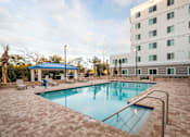 Thumbnail 6 of 11 - A swimming pool surrounded by a brick patio and a white building in the background.